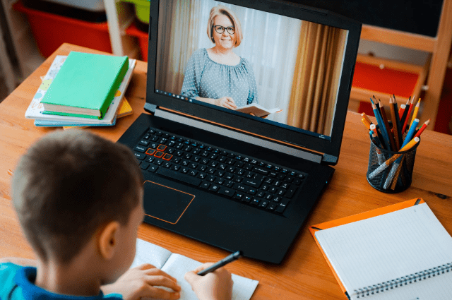 Female teacher on laptop screen smiling while young boy writes notes